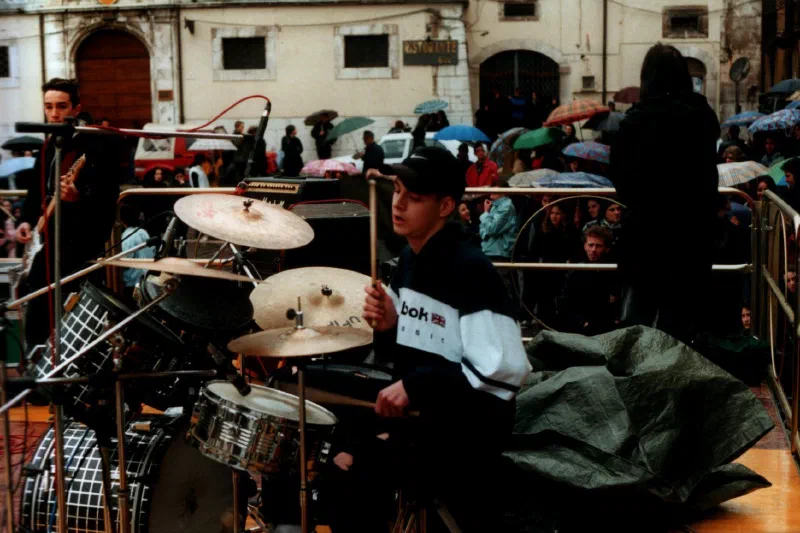 Fabio D’Amico playing drums at an outdoor gig in a town square, with the audience under umbrellas